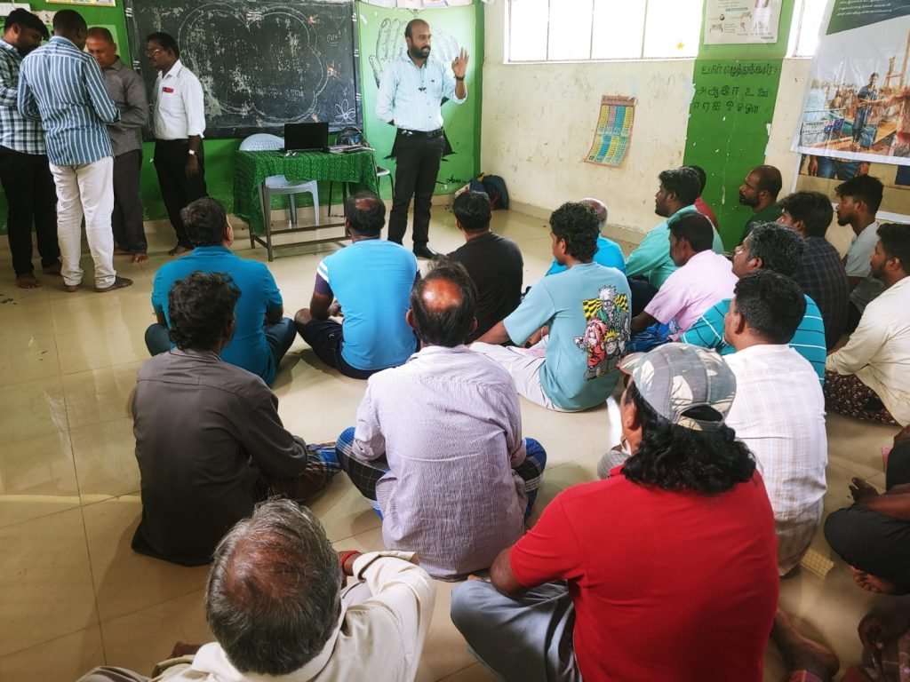 A speaker addressing a group of painters seated on the floor during a briefing session.