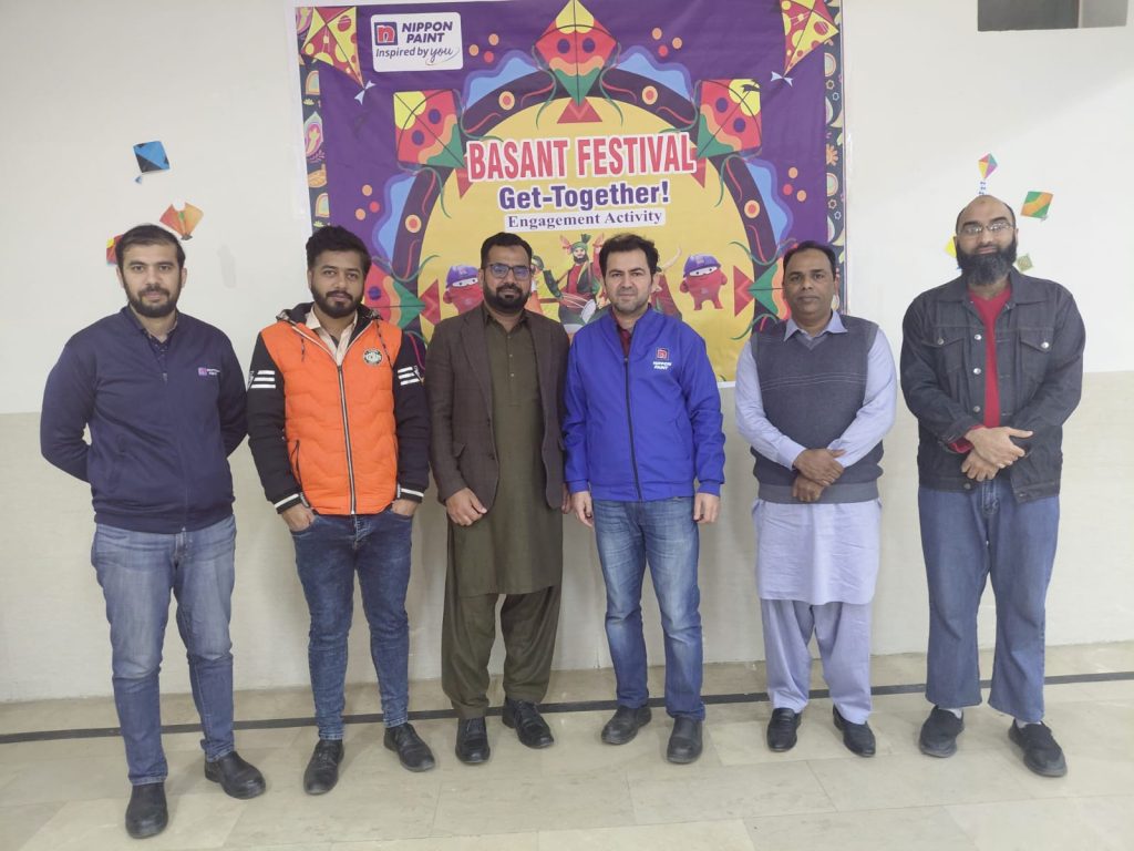 Six men standing in front of a colourful Basant Festival backdrop.