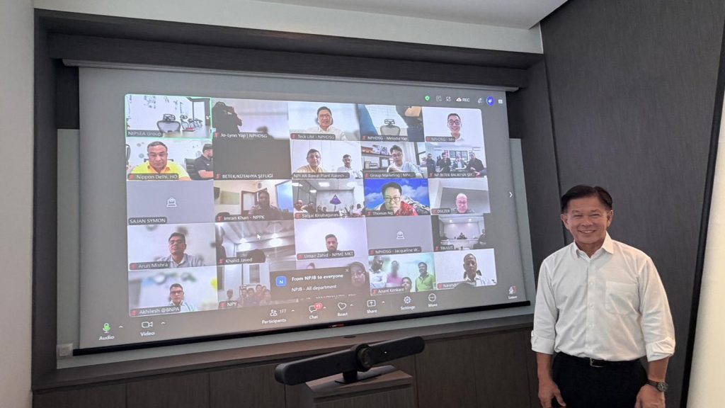 Group CEO Wee Siew Kim standing and smiling in front of a large screen displaying a global Zoom call with numerous regional participants.
