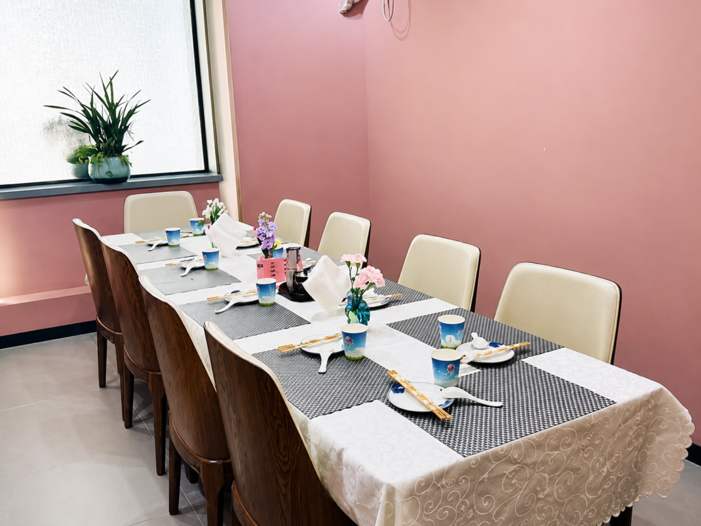A neatly set dining table with floral centrepieces inside a brightly lit, pink-walled private room.